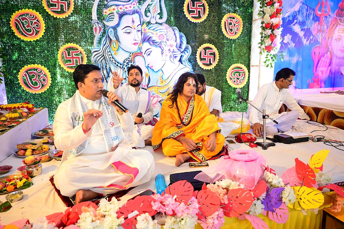 Acharya Pt. Binod Shastri seated with other priests before ceremonial materials and a decorated backdrop.